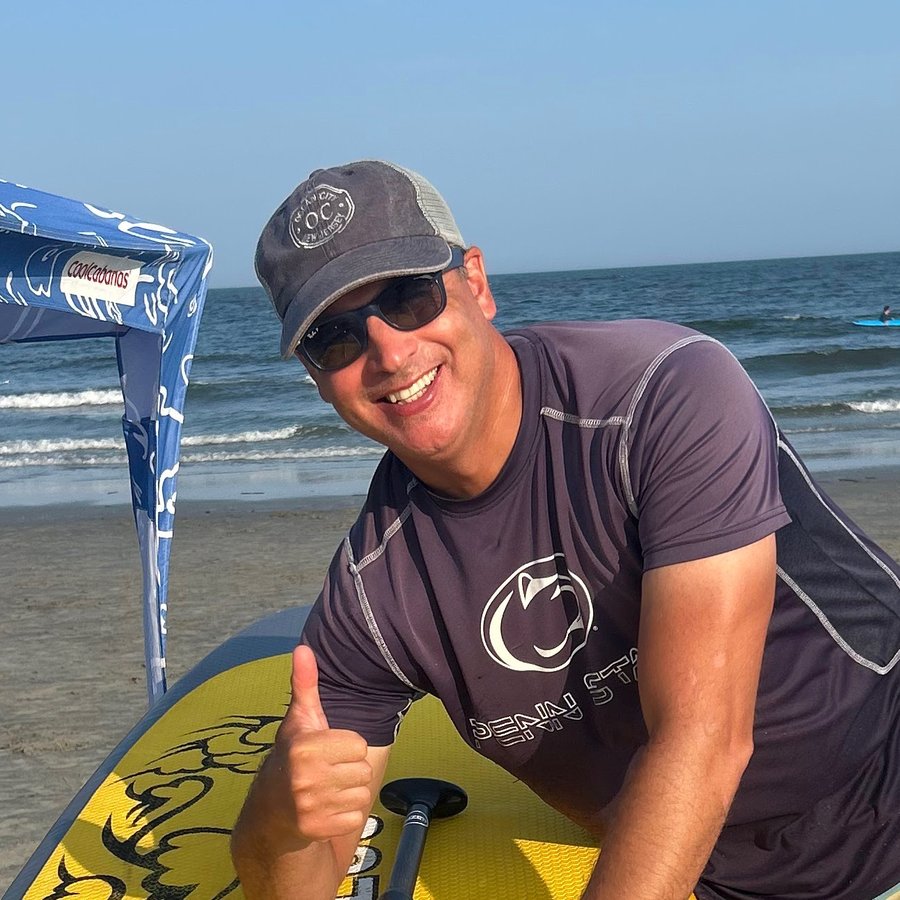 Kurt giving a thumbs up while leaning on his paddle board at the beach, wearing a Penn State shirt and an Ocean City cap